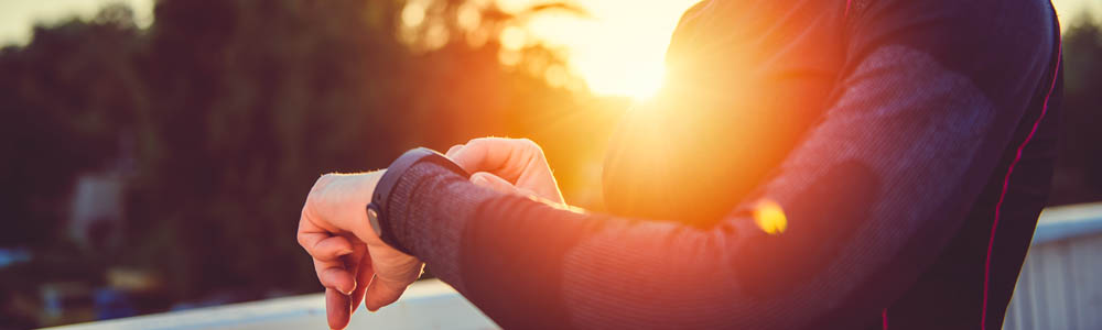 Close-up of woman's torso, she has one hand on her smart watch. Sun is bright in the background.